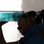 Frau schüttet sich an einem heißen Sommertag Wasser aus einer türkisfarbenen LumiVitae-Cellpower-Flasche über den Kopf. Wassertropfen spritzen, der Himmel ist wolkig dramatisch – sichtbare Erfrischung bei Hitze.
