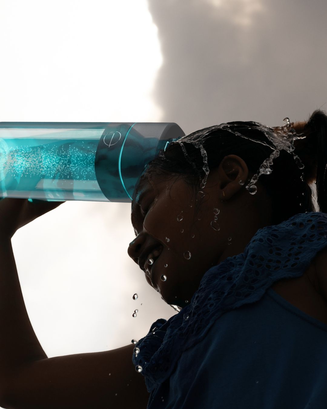 Frau schüttet sich an einem heißen Sommertag Wasser aus einer türkisfarbenen LumiVitae-Cellpower-Flasche über den Kopf. Wassertropfen spritzen, der Himmel ist wolkig dramatisch – sichtbare Erfrischung bei Hitze.