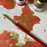 Abstraktes rot-goldenes Gemälde mit Pinsel und kleinen Farbfläschchen auf einem Arbeitstisch.