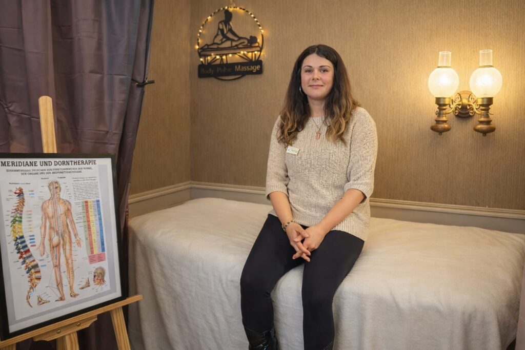 Anastazija Miskic sitzt auf einer Massageliege in ihrem Behandlungsraum und blickt in die Kamera.
